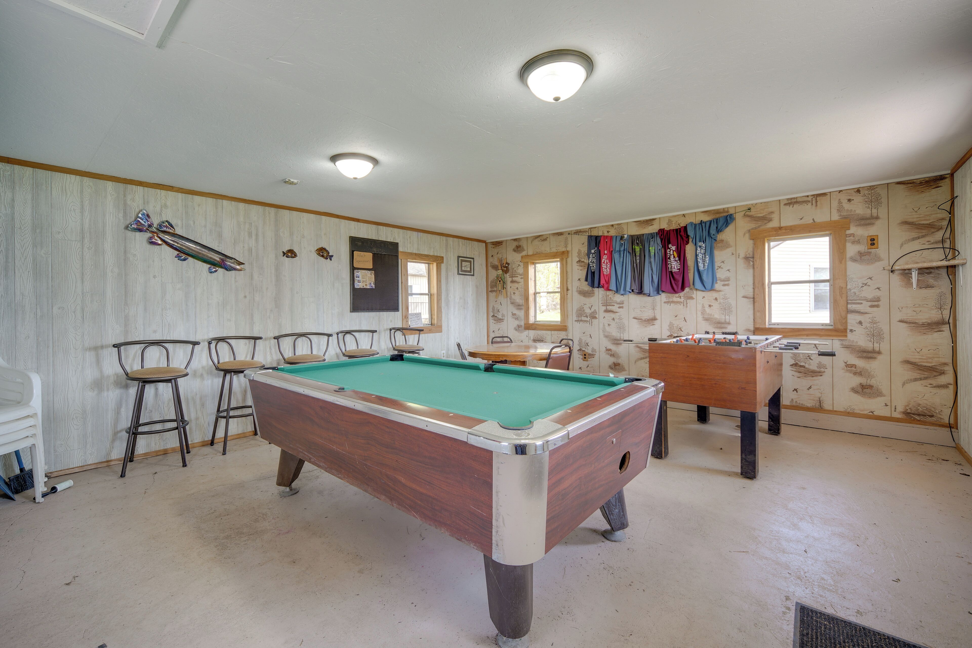 A pool table in a room with wooden paneling, a foosball table, and four bar stools.