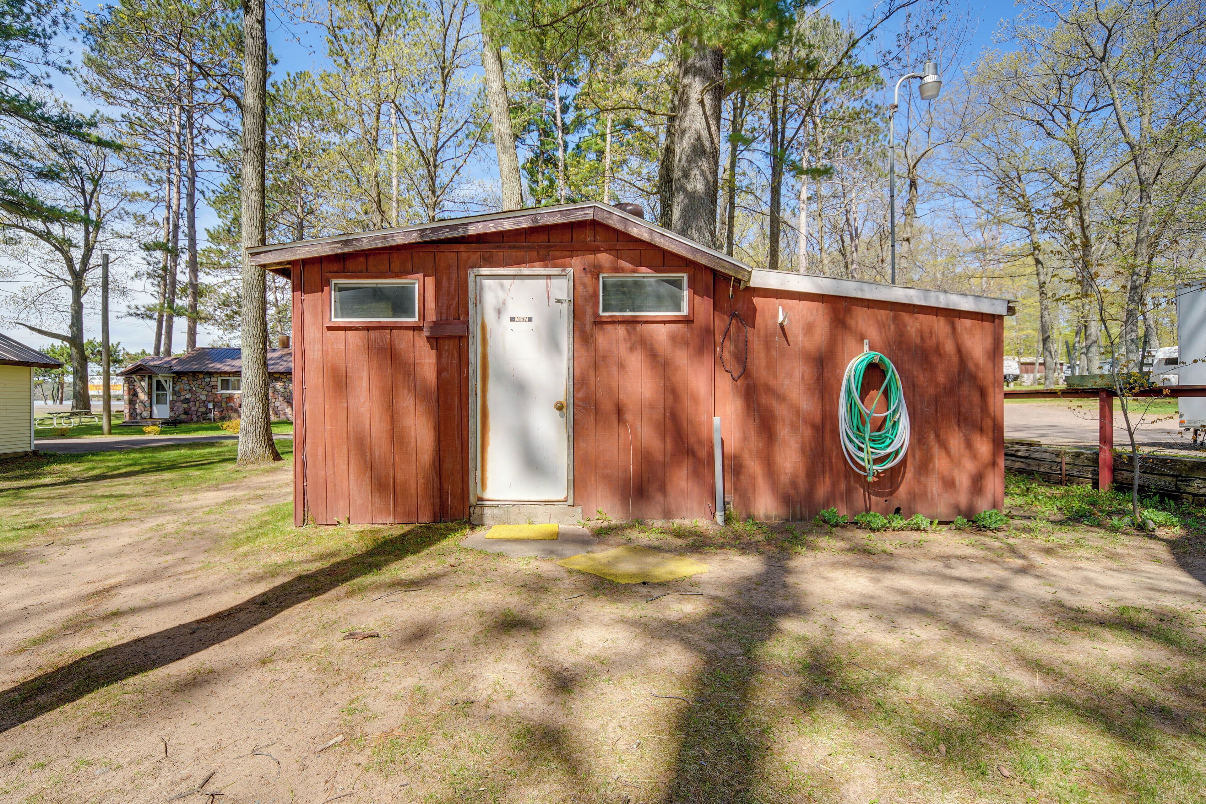 Une petite cabane en bois, dotée d’une porte et de fenêtres, entourée d’arbres et d’une zone herbeuse.
