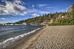 A beach with a wooden pier extending into calm waters, surrounded by dense forest and a clear sky. - Charming Downtown Coeur D'alene Home With Yard! (Coeur d'Alene)