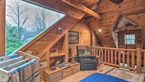 Ein gemütliches Schlafzimmer unter dem Dach mit geneigter Decke, Holzbalken, Deckenventilator, großem Fenster und Blick ins Freie.