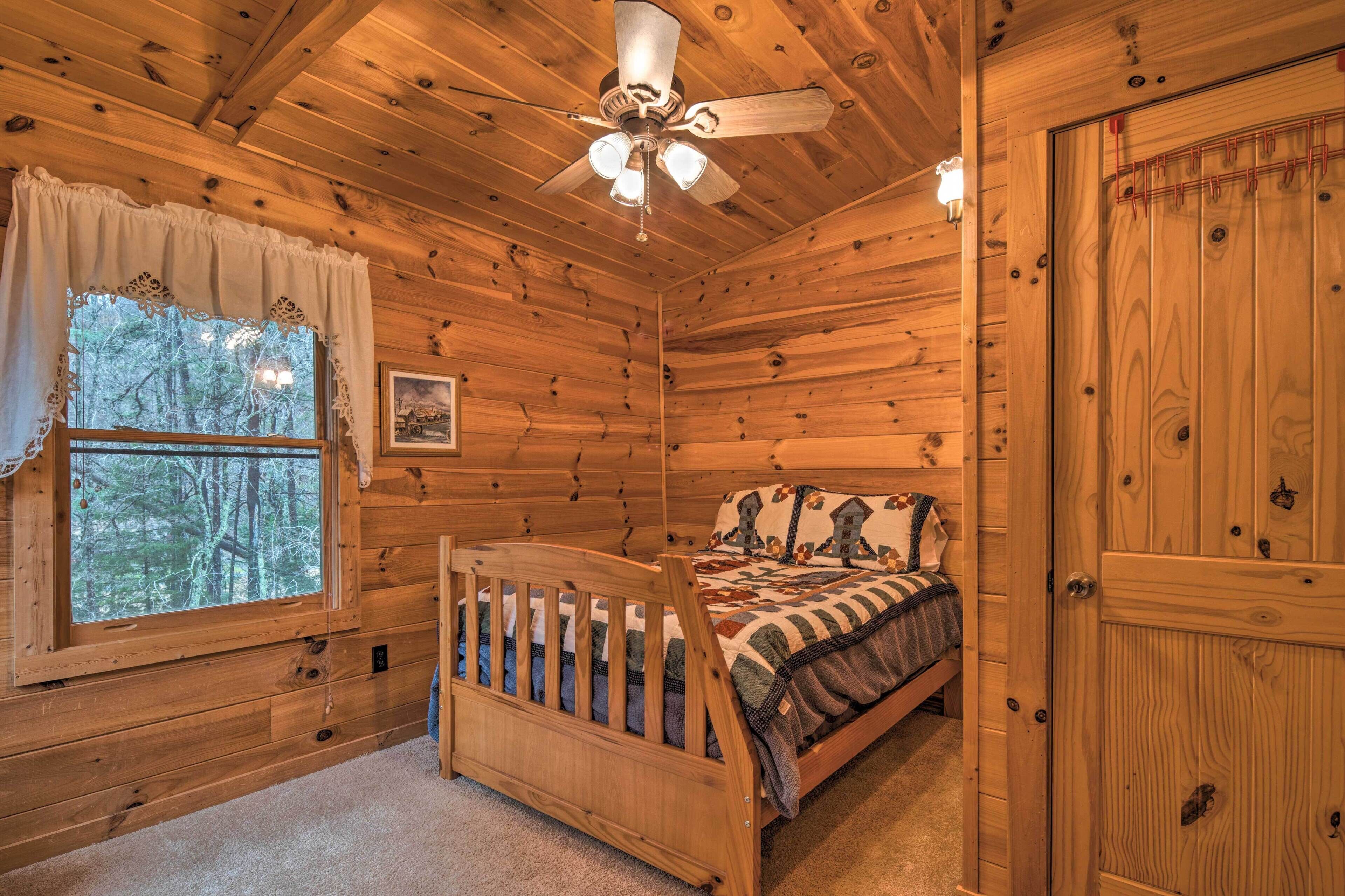 Un dormitorio de cabaña de madera con cama, cuna, ventilador de techo, ventana con cortinas y puerta.