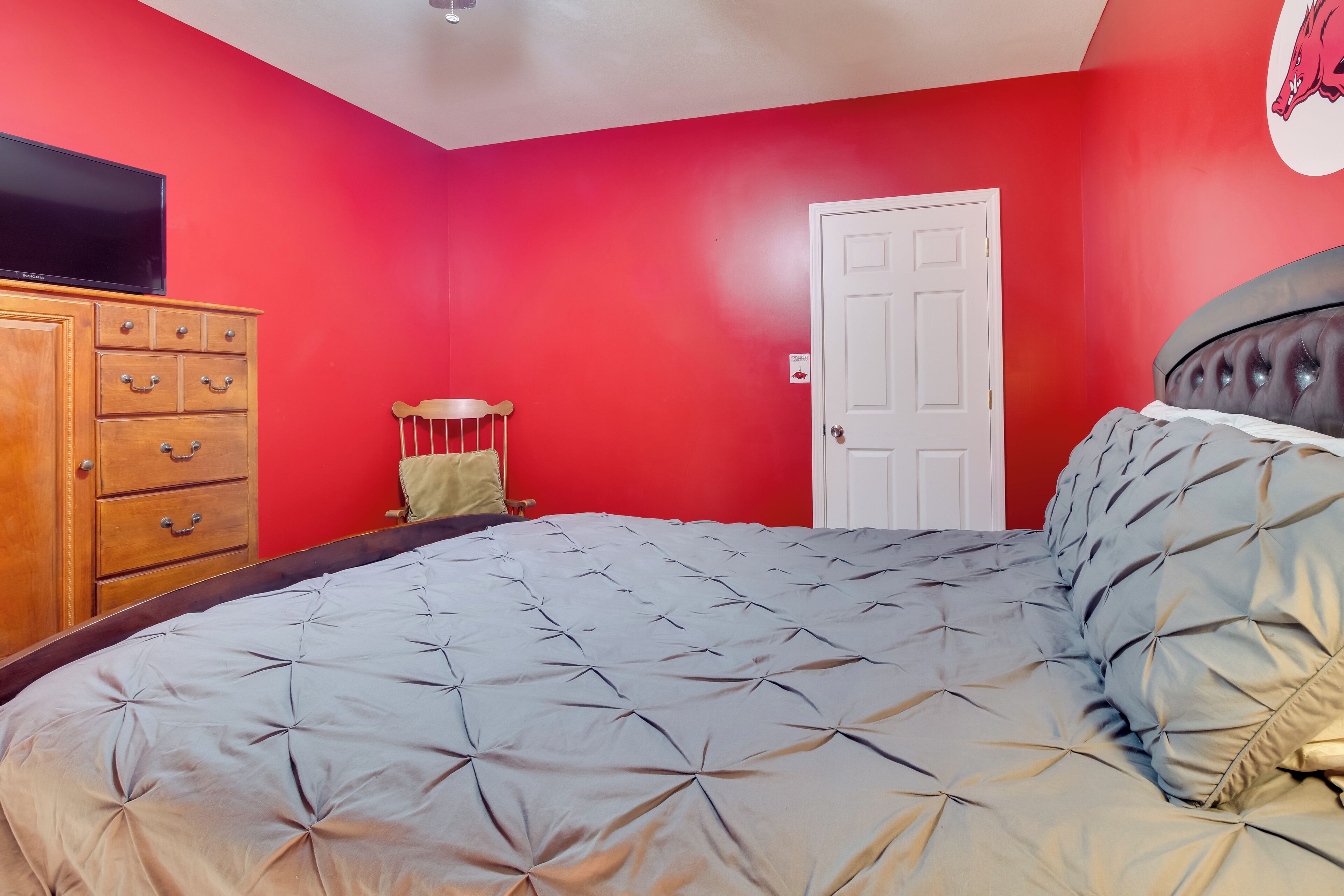 A bedroom with a red wall, a wooden dresser, a bed with a grey quilt, and a white door.