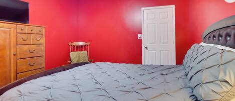 A bedroom with a red wall, a wooden dresser, a bed with a grey quilt, and a white door.