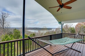 A wooden deck with a hammock and a ceiling fan, overlooking a lake and forested hills. - Spacious Norfork Lake Home: Game Room + Views! (Elizabeth)