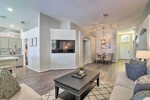 A modern living room with a flat-screen TV, a coffee table, and a sofa. There is a kitchen area with cabinets and a dining table in the background. - 5 Mi to Disney World: Retreat w/ Pool & Lanai! (Kissimmee)