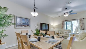A dining area with a wooden table set for four, surrounded by wooden chairs. There is a potted plant on the left and a painting on the wall. A ceiling fan and a TV are visible in the background. - Kissimmee Retreat w/ Pool Access < 4 Mi to Disney! (Kissimmee)