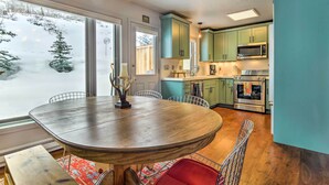 Een eetruimte met een houten tafel en stoelen, een uitzicht op een besneeerd landschap door grote ramen, en een keuken met blauwgroene kastjes en moderne apparatuur.