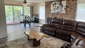 A spacious living room with a leather sofa, a coffee table with a natural wood top, and a large abstract painting on the brick wall.