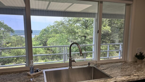 A kitchen with a stainless steel sink, a marble countertop, and a view of a balcony with trees and a body of water.
