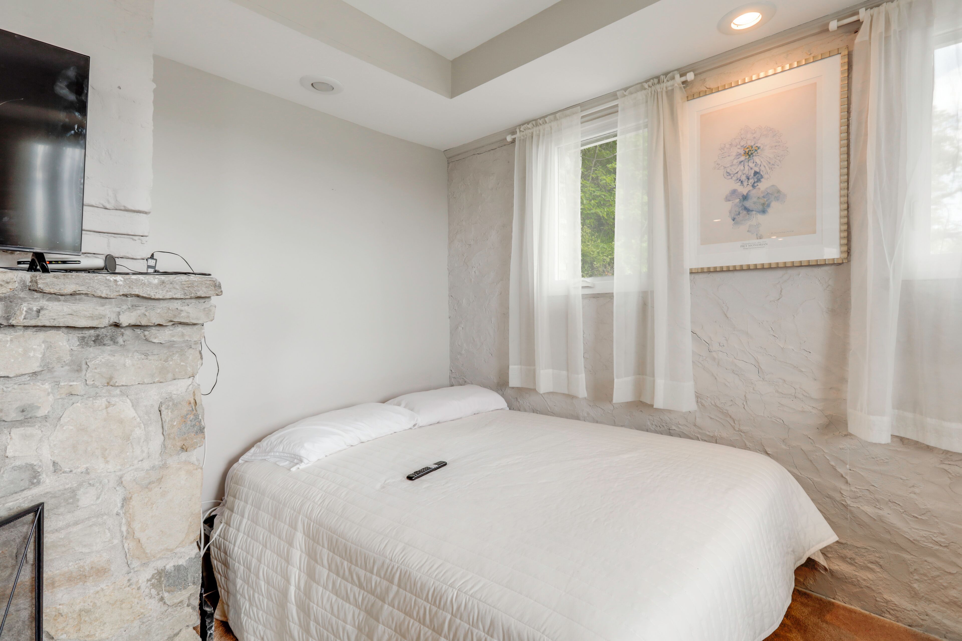 A bedroom with a stone fireplace, a bed with white bedding, a remote control on the bed, a framed artwork above the bed, and a window with sheer curtains.