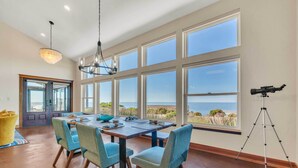 Un comedor con una mesa grande y sillas, ventanas amplias con vista al océano, y un trípode en el suelo.