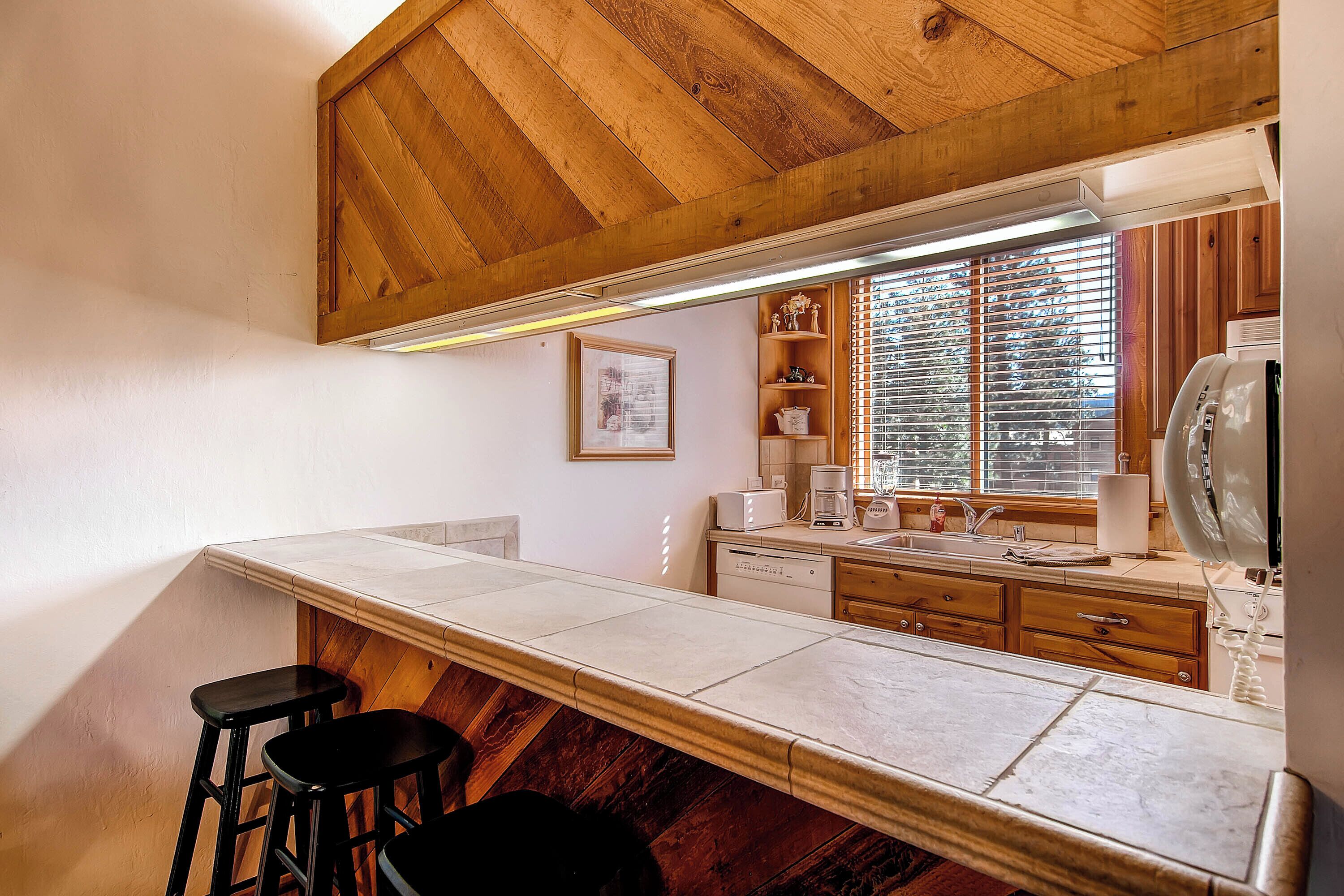 A kitchen with a wooden ceiling, a long counter, and a window with blinds.