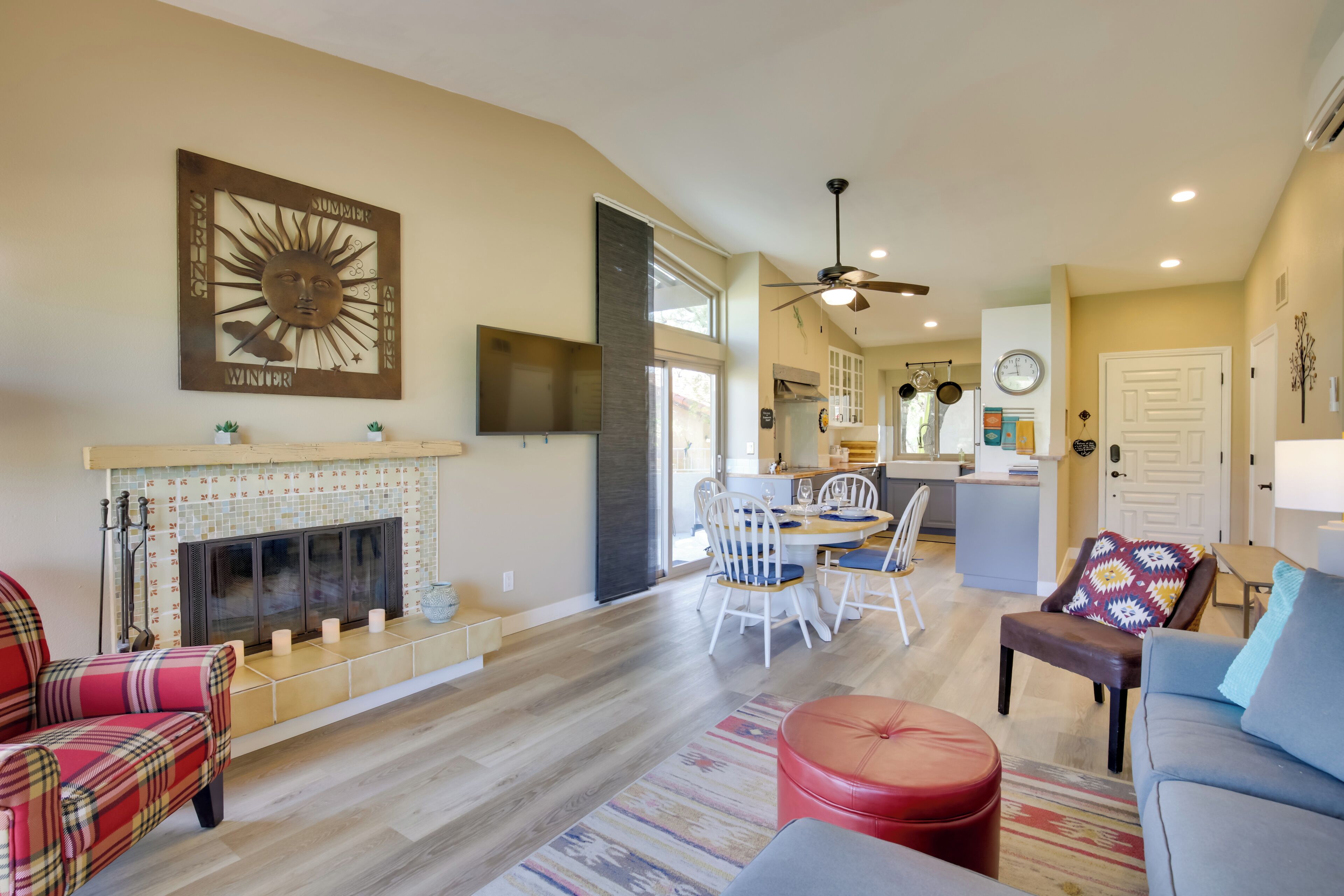 A spacious living area with a fireplace, a dining table with chairs, a kitchen in the background, and a ceiling fan.
