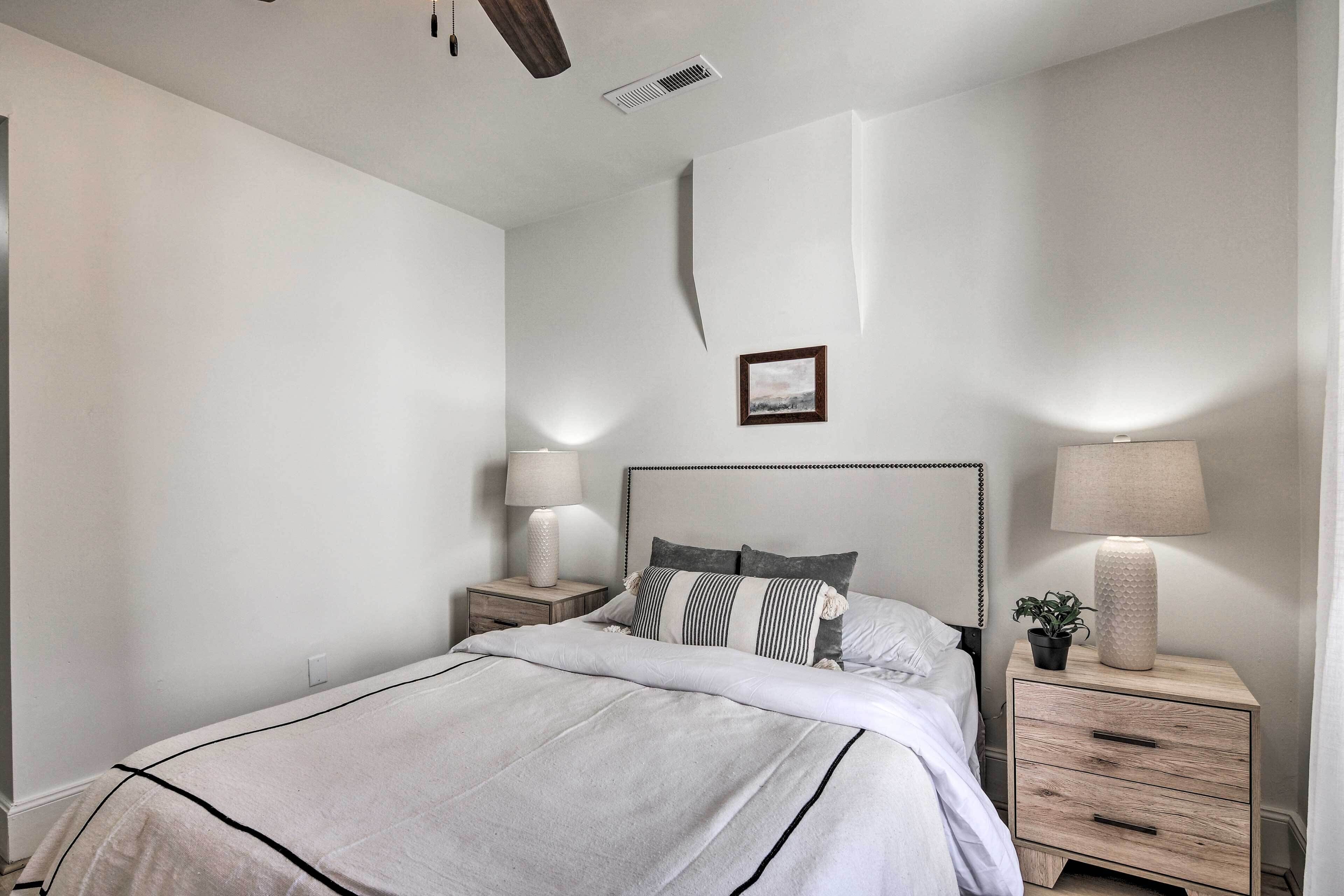 A bedroom with a bed, two bedside tables, a nightstand, a lamp, a framed picture, and a potted plant.