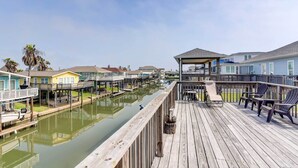 Eine hölzerne Pier mit Stühlen und einem überdachten Bereich, die zu einem Kanal mit Hausbohlenhäusern führt.