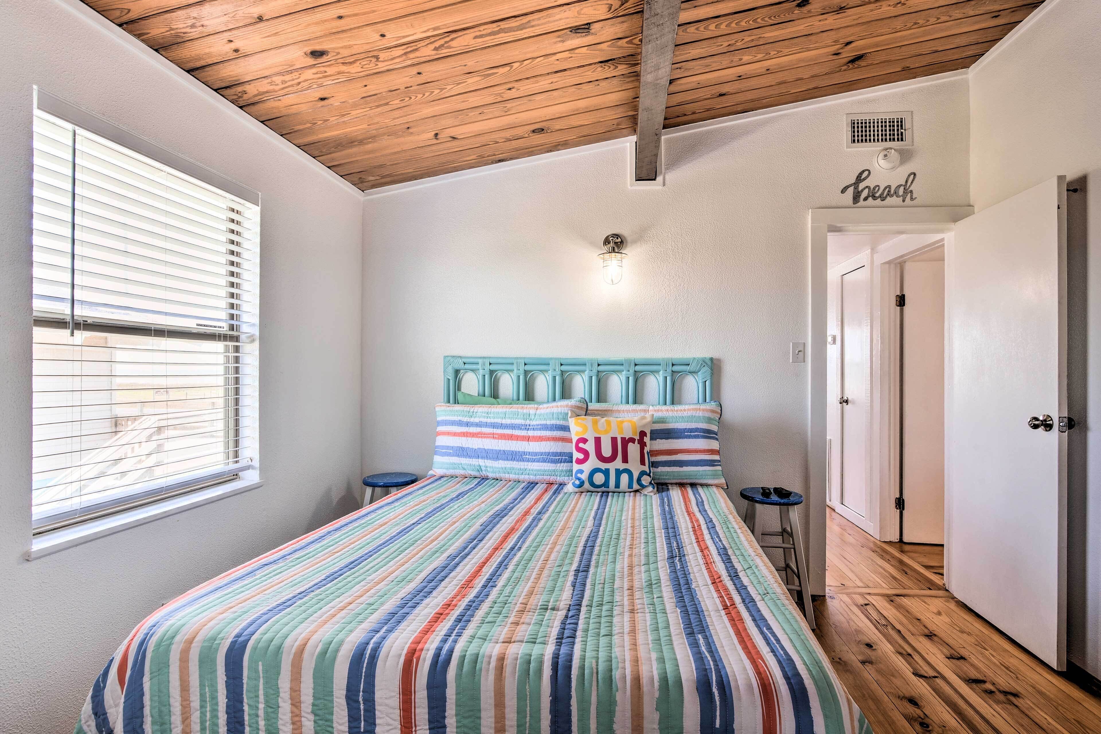 A bedroom with a wooden ceiling, a bed with a colorful striped bedspread, a window with blinds, and a door labeled "Beach".
