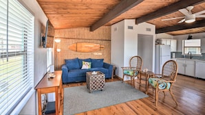 A cozy living area with a wooden ceiling, a blue sofa, a small table, wicker chairs, and a kitchen in the background. - Gulf Coast Views: Stilted Retreat in Galveston! (Galveston)