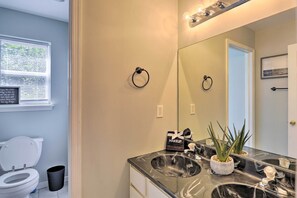 Une salle de bain avec des toilettes blanches, une poubelle noire, un lavabo avec un miroir et un pot d’aloès.