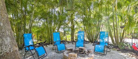 A seating area with four blue chairs and a fire pit surrounded by gravel, set in a tropical garden with palm trees.