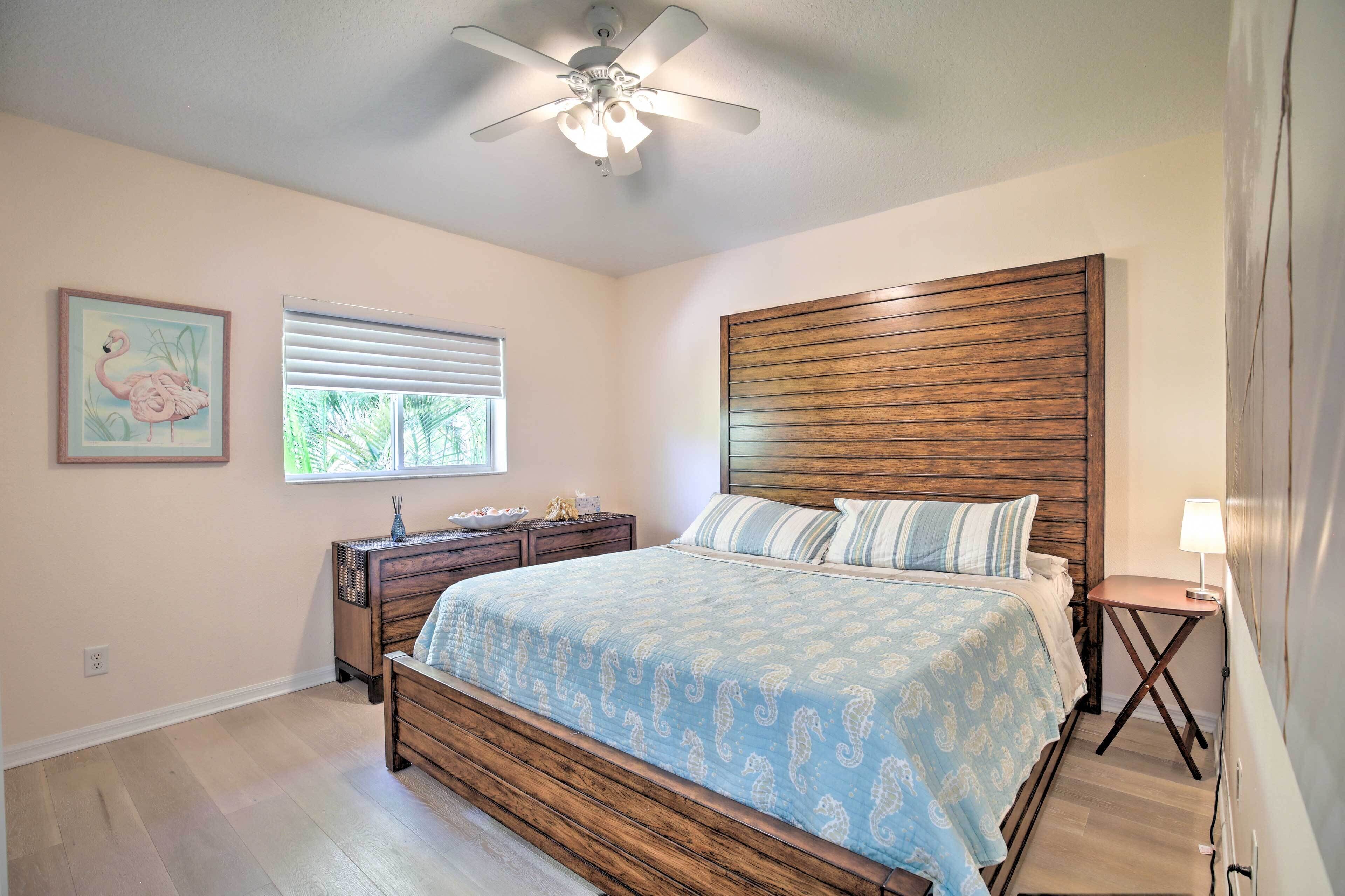 A bedroom with a wooden headboard, a bed with a blue patterned bedspread, a nightstand, a small table with a lamp, and a ceiling fan.