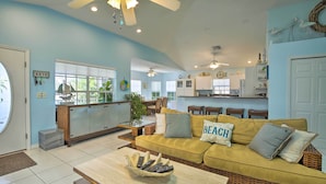 A spacious living room with a yellow sofa, a coffee table with remote controls, a ceiling fan, and a kitchen in the background. - Indian Rocks Beach Oasis - 2 Blocks to Beach! (Indian Rocks Beach)