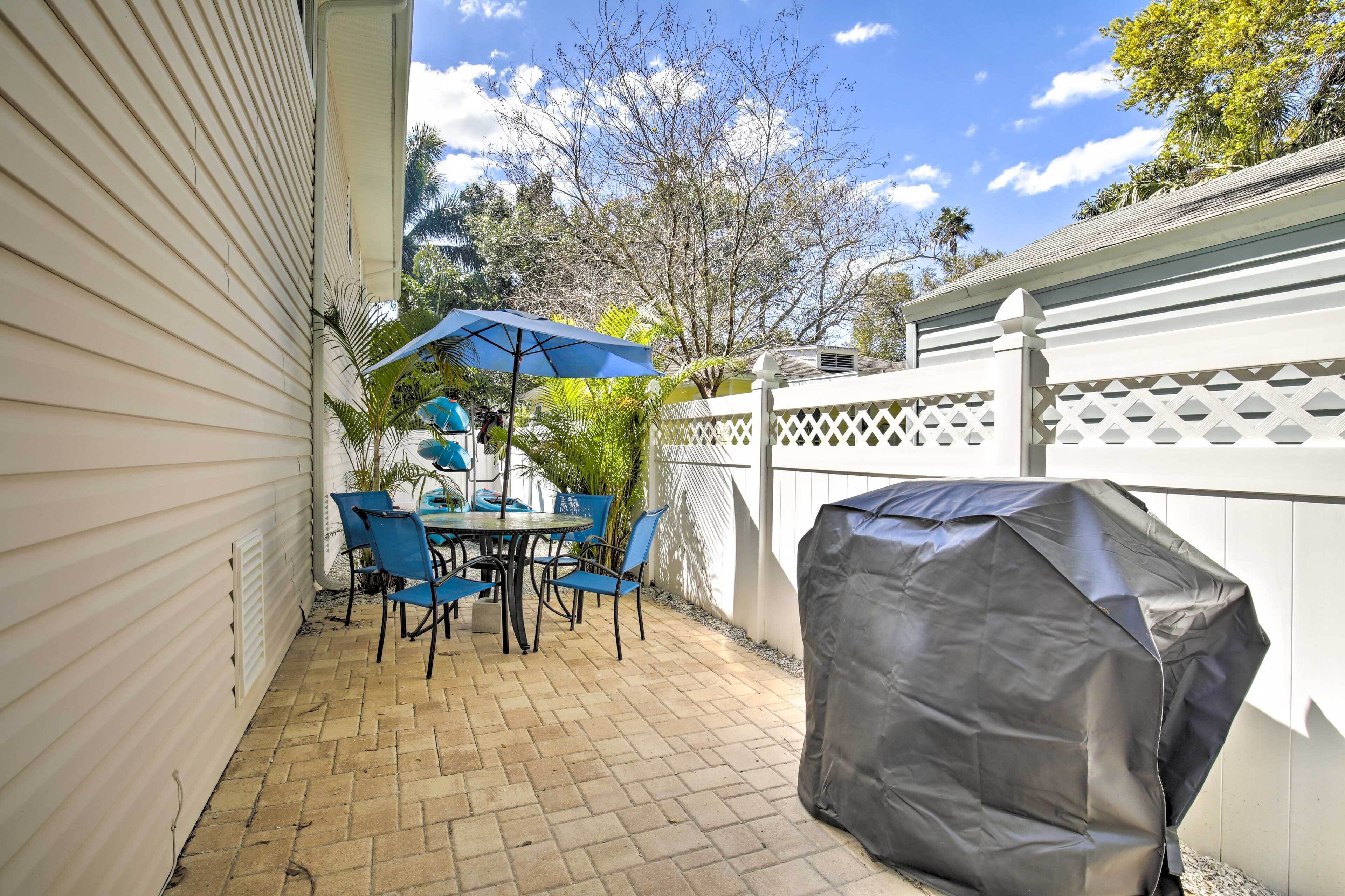 A patio with a table and chairs, a covered grill, and a white fence.