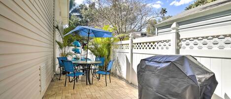 A patio with a table and chairs, a covered grill, and a white fence.