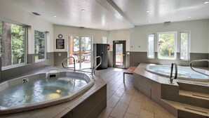 Un espace spa moderne doté de deux bains à remous, d’un banc en bois et de grandes fenêtres offrant une vue sur l’extérieur.