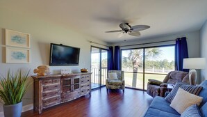 A spacious living room with a large TV, wooden furniture, a ceiling fan, and a view through large windows.