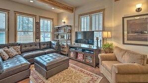 A living room with a sectional sofa, ottoman, and a TV stand with shelves and a flat-screen TV. - Updated Townhome w/ Hot Tub - Walk to Downtown! (Red Lodge)