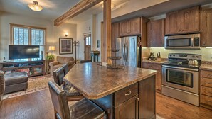 A spacious kitchen with wooden cabinets, a large island, stainless steel appliances, and a cozy living area with leather seating. - Updated Townhome w/ Hot Tub - Walk to Downtown! (Red Lodge)