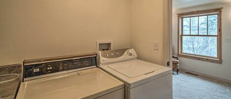 A laundry room with a front-loading washer and dryer, a small shelf with toiletries, and a window with a view of trees.