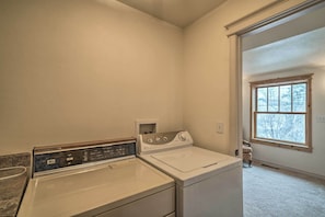 A laundry room with a front-loading washer and dryer, a small shelf with toiletries, and a window with a view of trees.