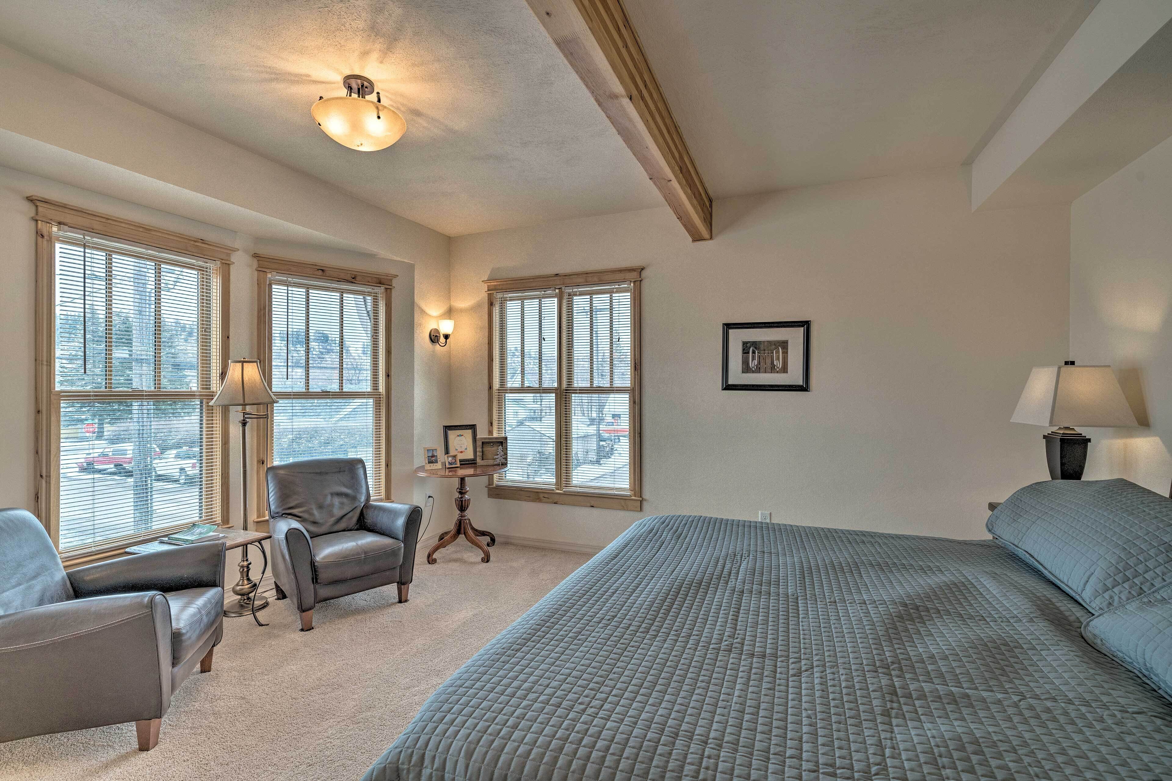 A bedroom with a large bed, two armchairs, a small table, and a window with blinds.