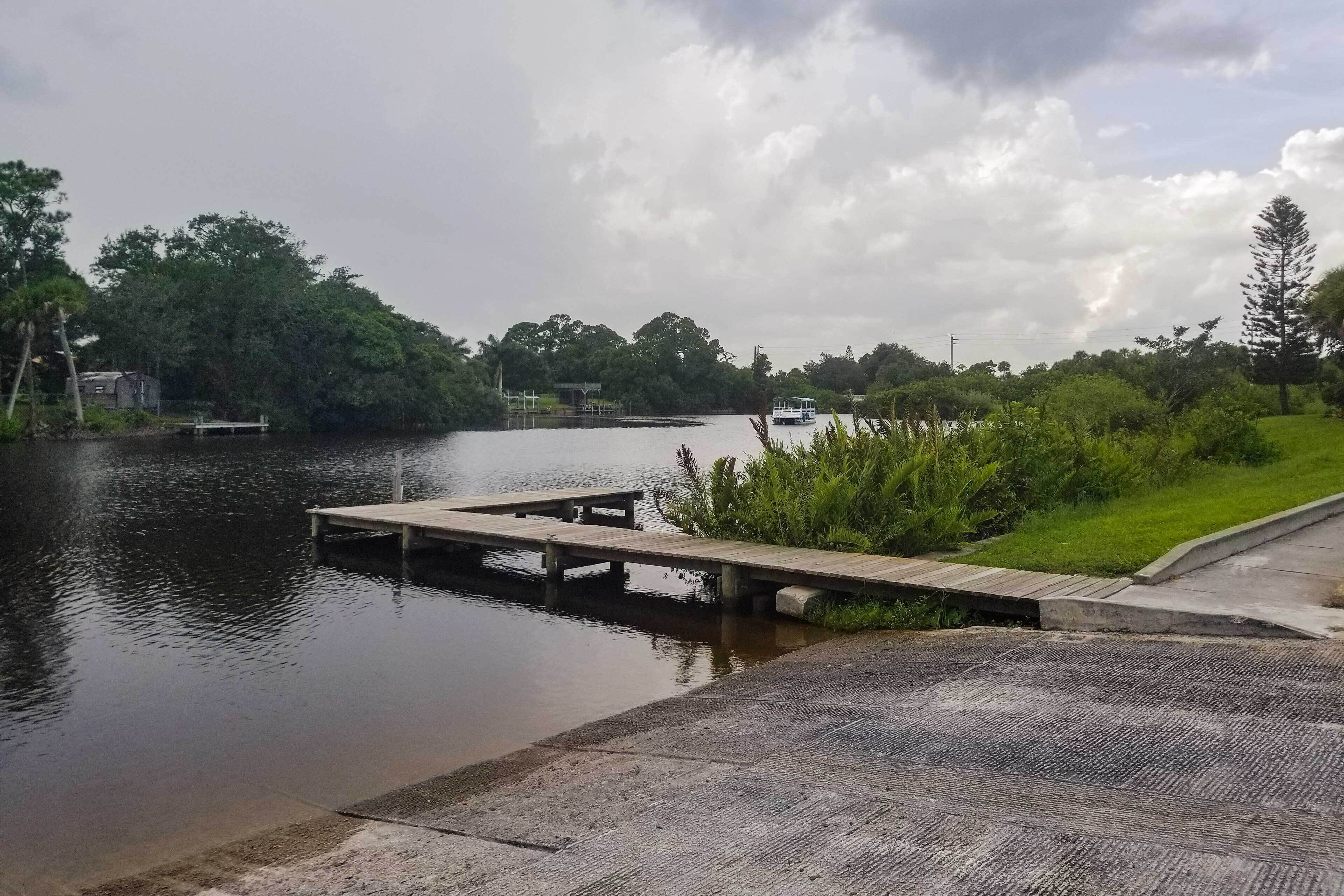 Un lago tranquilo con un muelle de madera que se adentra en el agua, rodeado de exuberante vegetación y un cielo nublado.