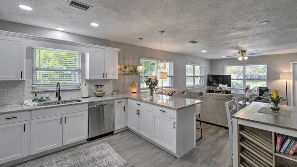 A modern kitchen with white cabinets, granite countertops, and a central island. There are pendant lights, a built-in oven, and a large window providing natural light. - Bright Venice Getaway < 1 Mi to Historic Dtwn (Venice)