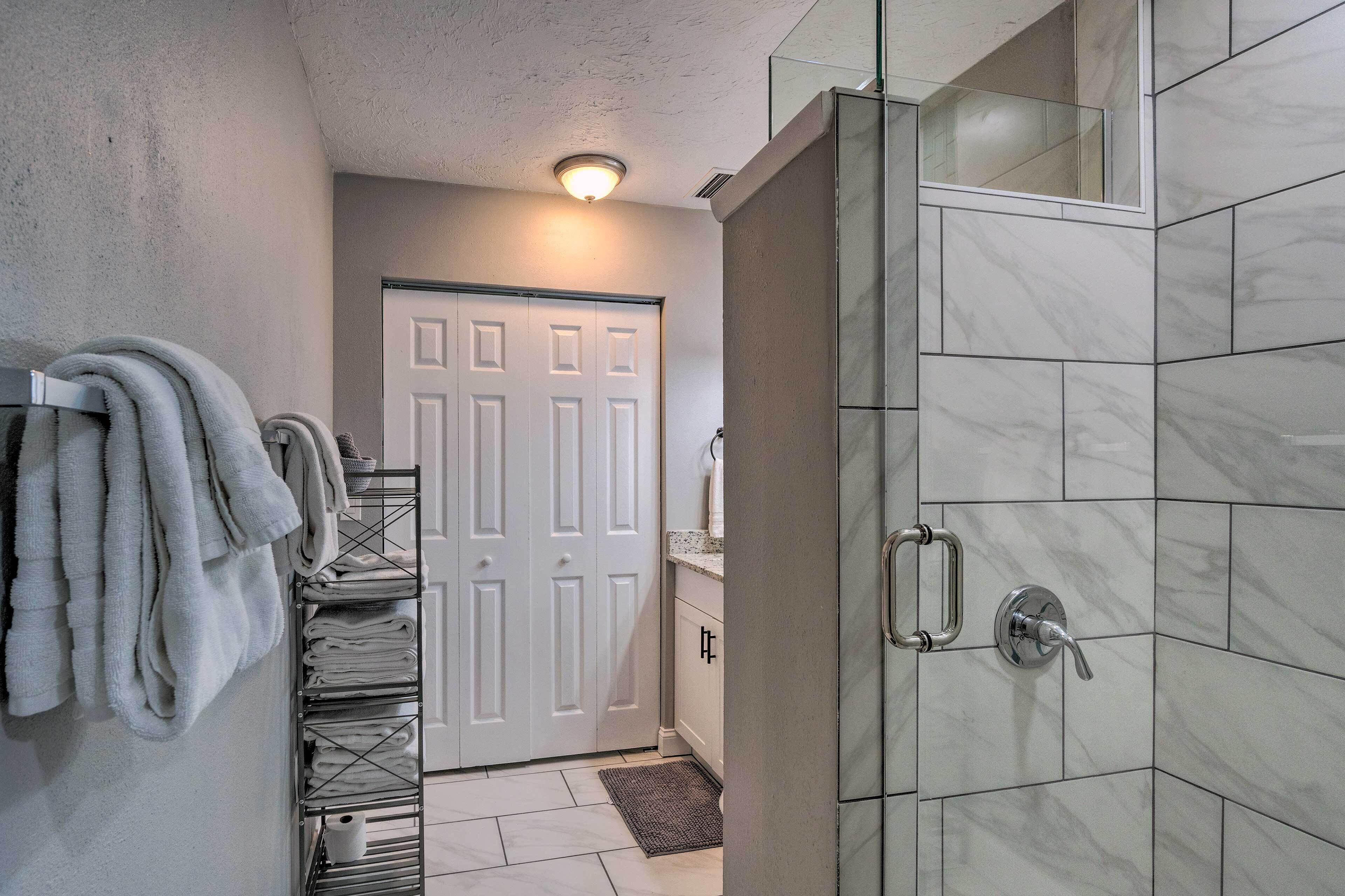 A bathroom with a glass shower enclosure, a towel rack with folded towels, and a white door in the background.