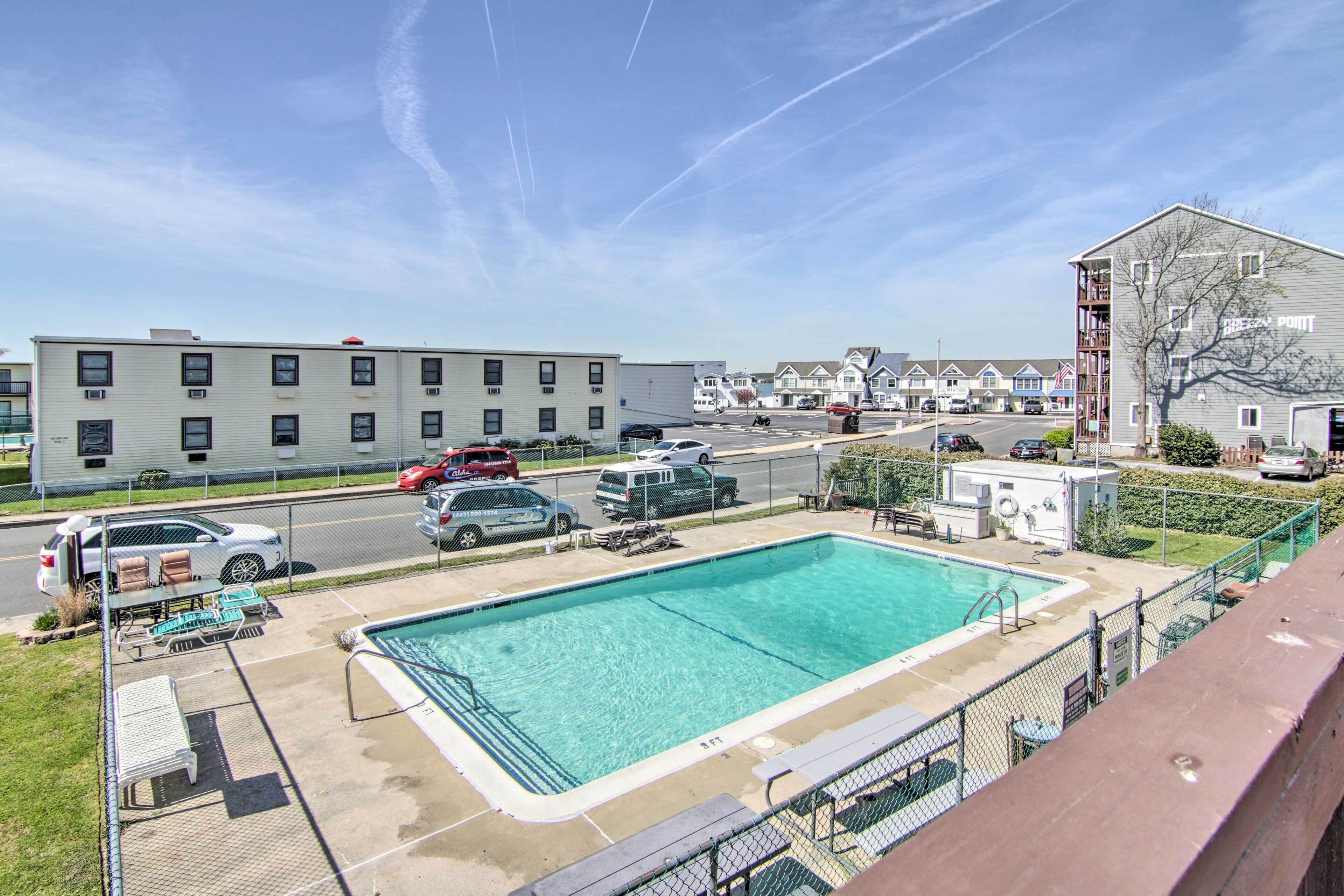 A swimming pool with a fenced deck, a parking lot with several cars, and a row of buildings in the background.
