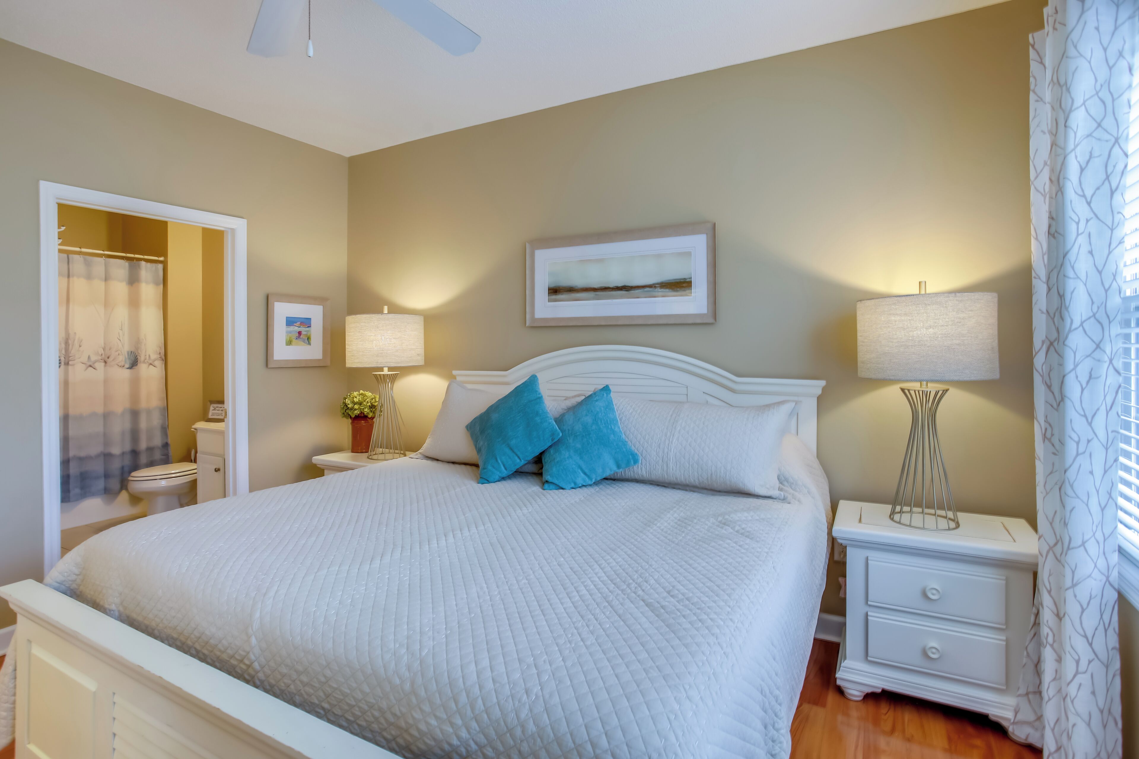 Une chambre à coucher avec un grand lit, deux tables de chevet, une commode, un ventilateur de plafond et une fenêtre avec des rideaux.