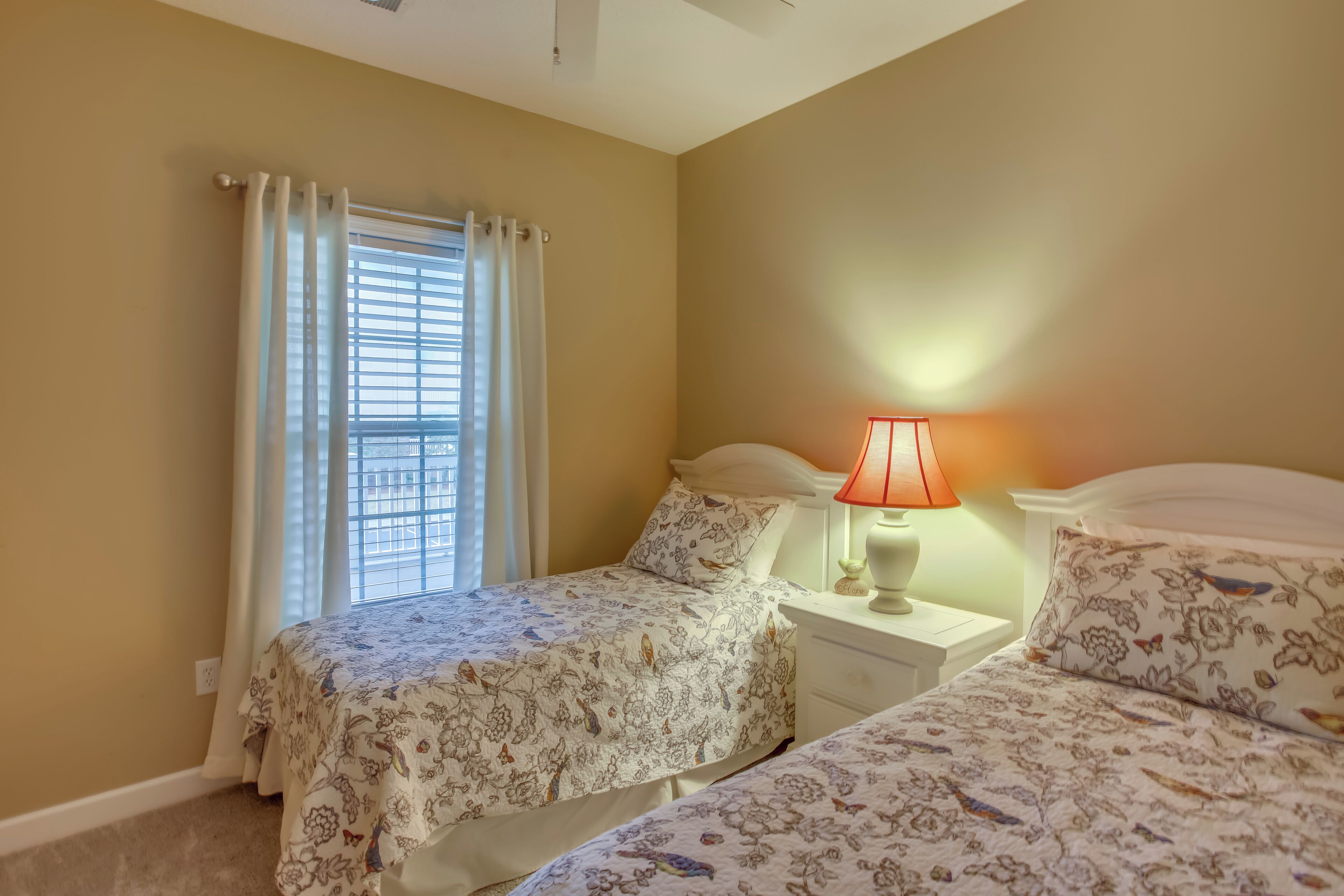 Une chambre avec deux lits, une fenêtre avec des rideaux, une table de chevet avec une lampe et un mur avec un interrupteur.
