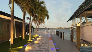 A waterfront walkway with palm trees, a boat dock, and a building with a glass facade. - Matlacha Waterfront Retreat: Dock & Hot Tub! (Matlacha)