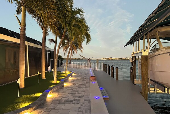 A waterfront walkway with palm trees, a boat dock, and a building with a glass facade.