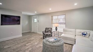 A spacious living room with a grey ottoman, a white sofa with a blue crab pillow, a patterned armchair, a large painting on the wall, and a window with blinds. - Matlacha Waterfront Retreat: Dock & Hot Tub! (Matlacha)