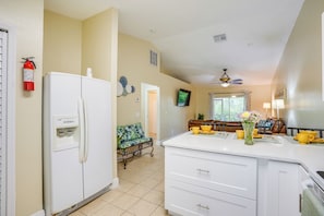A kitchen with a white refrigerator, a coffee machine, and a sink. There is a window with curtains and a ceiling fan.