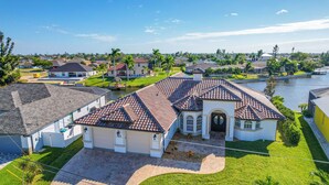 A residential house with a tiled roof, a well-maintained lawn, and a body of water in the background. - On Canal: Luxe Cape Coral Oasis w/ Pool & Lanai! (Cape Coral)