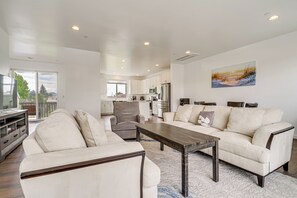 A modern living room with a sofa, armchair, and coffee table, a kitchen in the background, and a painting on the wall.