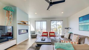 A modern living room with a flat-screen TV, a ceiling fan, a large window, and a sofa with patterned cushions. - Scenic Depoe Bay Townhome w/ Deck < 1 Mi to Parks! (Depoe Bay)