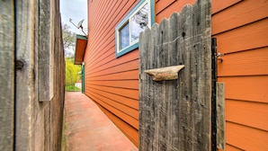 Un étroit sentier mène à une porte en bois sur laquelle est apposée une pancarte indiquant « Entrée ». De chaque côté de la porte, on aperçoit un bardage en bois et une fenêtre dotée d’un cadre bleu.
