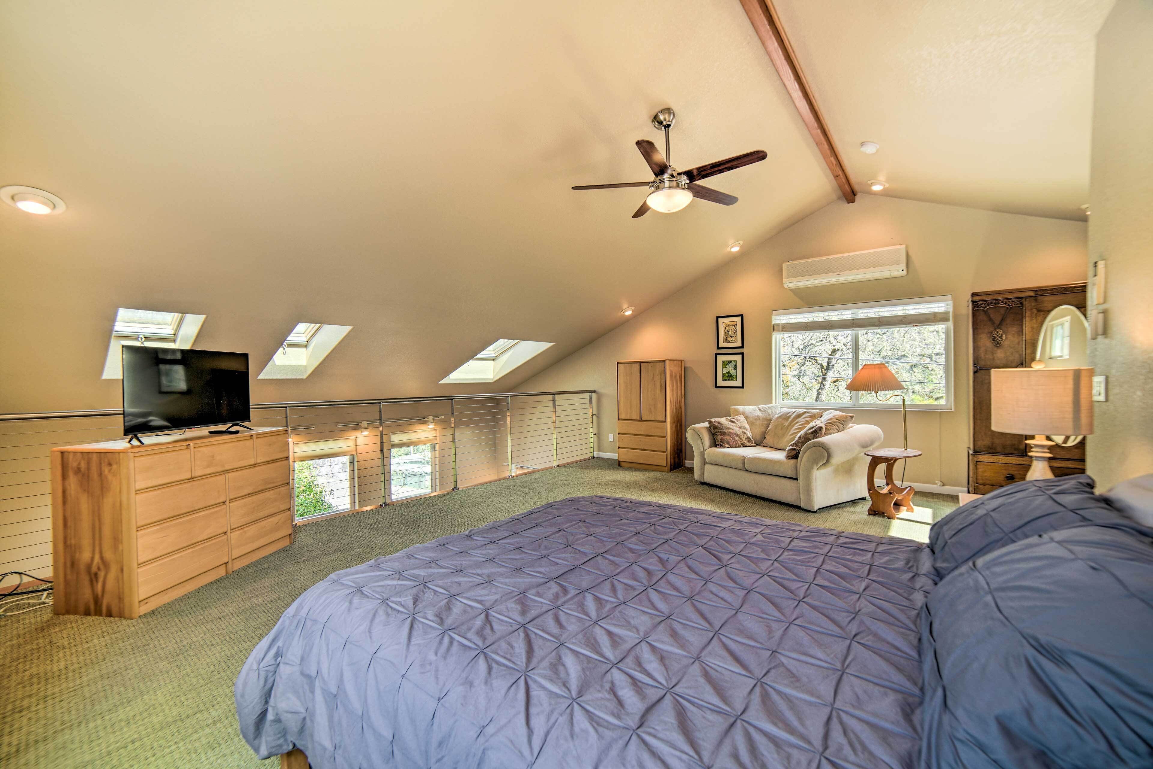 A bedroom with a large bed, a ceiling fan, a TV on a wooden dresser, a window with a view, and a small table with a lamp.