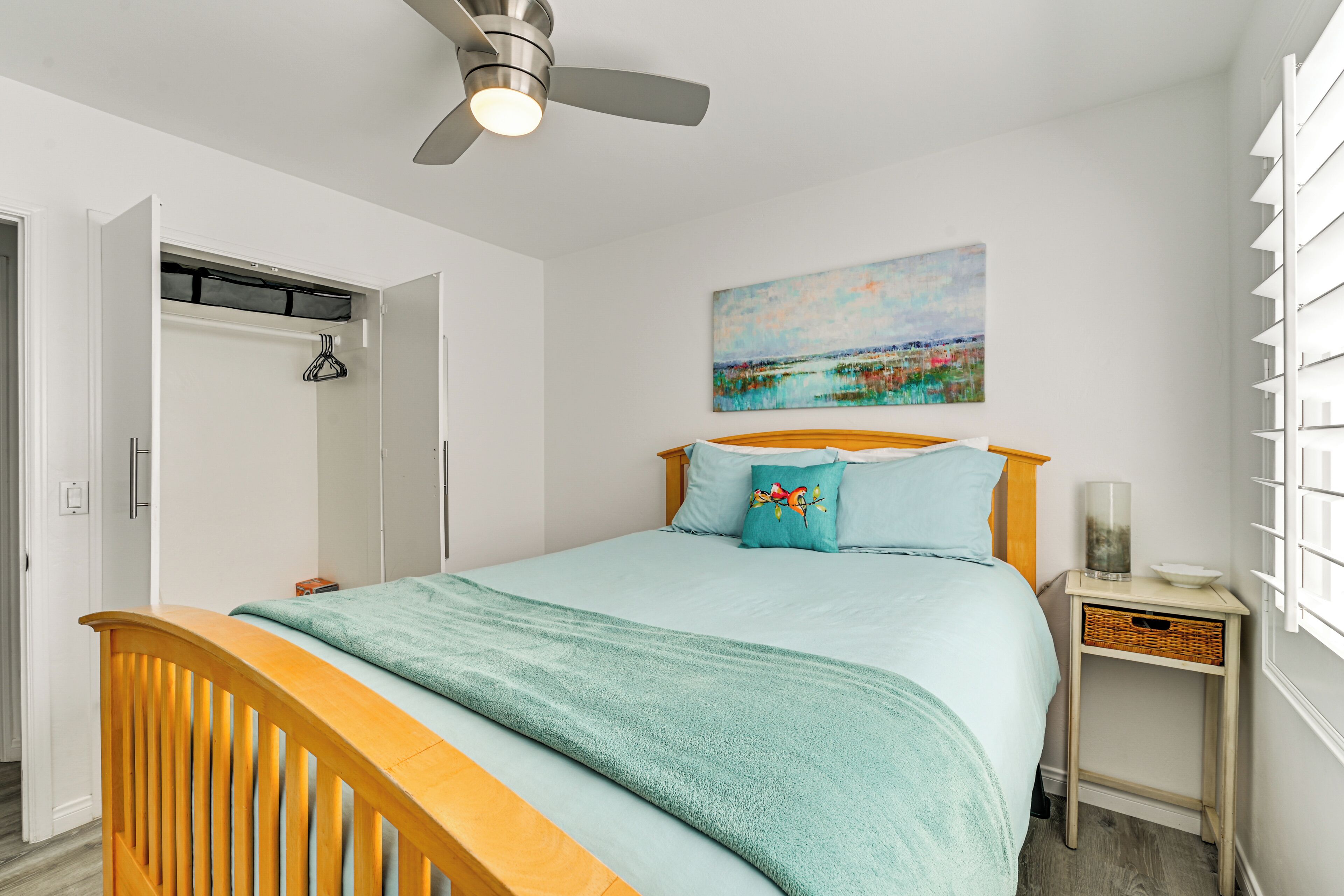 Un dormitorio con cama, ventilador de techo, mesita de noche, estante en la pared y un cuadro.
